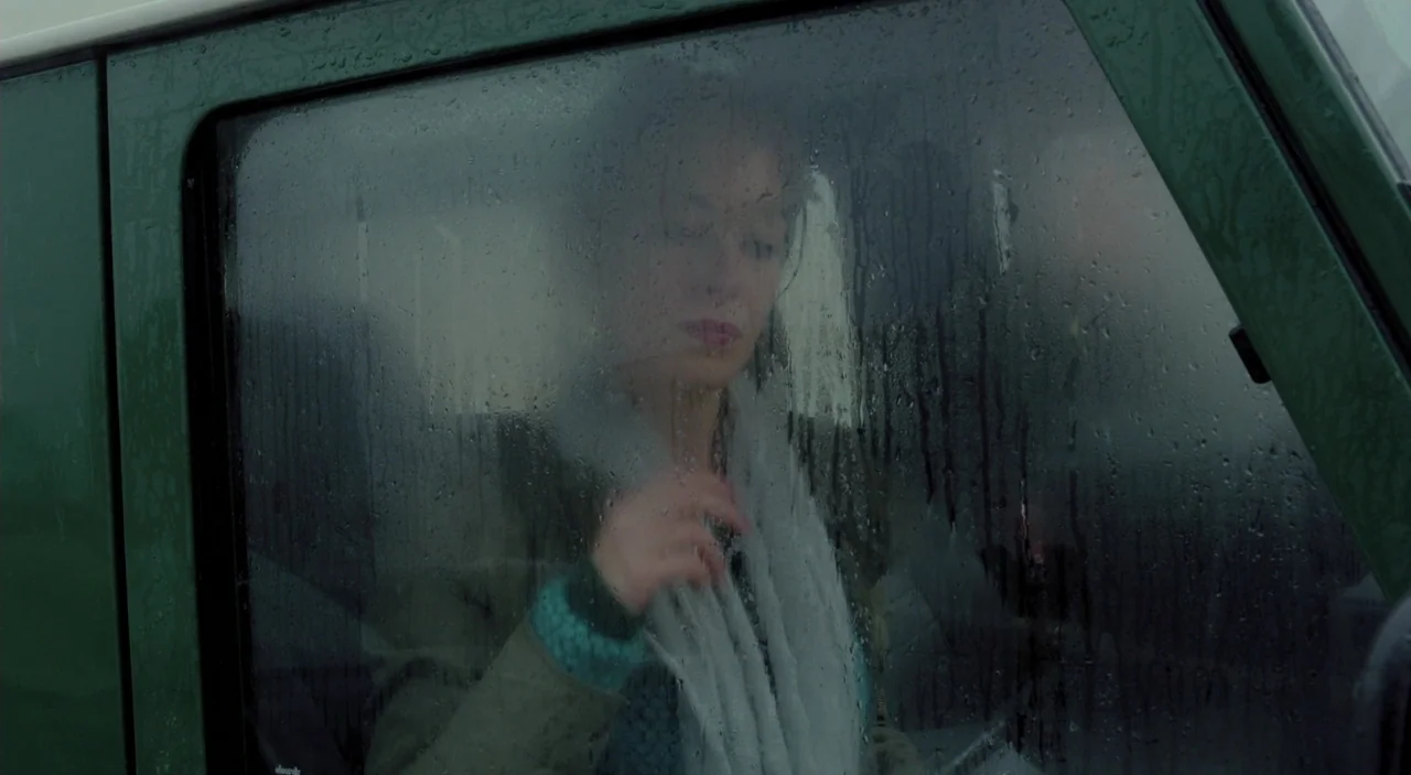 Cosmos (2015) movie still: medium — The interior of a vehicle seen through a wet, misted-over passenger window. A woman sits in the seat and adjusts…