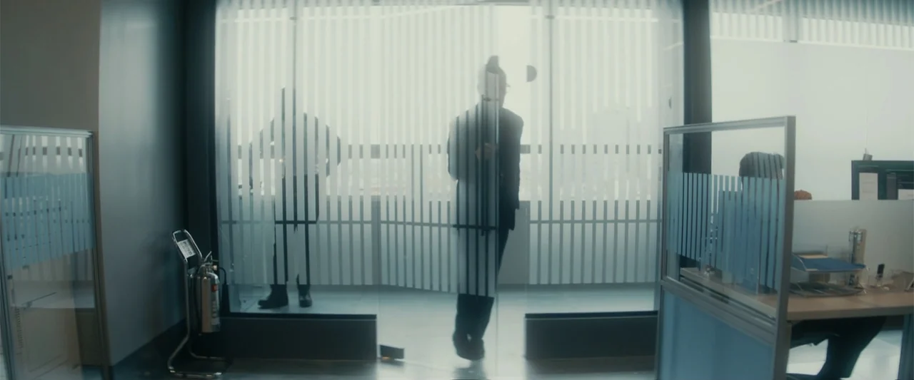 Black Bag (2025) movie still: wide — A modern corporate workspace features glass partitions with vertical frosted blind patterns. A man stands in…