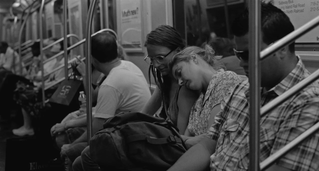Frances Ha (2012) movie still: medium — The interior of a commuter subway car with metal poles and overhead advertisements. A woman sleeps with her…