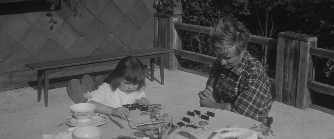 Jules and Jim (1962) movie still: medium — A wooden patio features a round table set for activity with wooden benches nearby. A young girl and an…