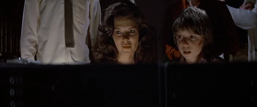 Poltergeist movie still: medium — A dark space with electronic consoles illuminated by the glow of monitors. A woman and a young boy look forward…