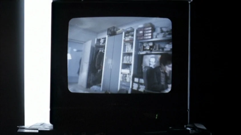 Benny’s Video (1992) movie still: medium — A dark room features a glowing television screen showing an interior space. A man is seated at a desk…