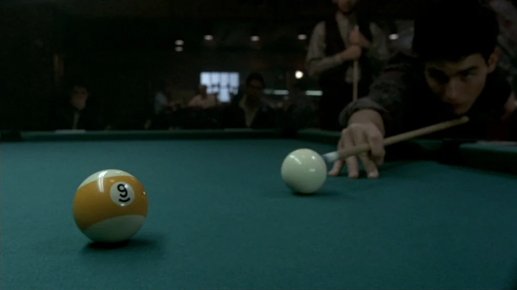 The Colour of Money (1986) movie still: medium — A dimly lit billiard hall features a green felt table as the central focus. A young man leans…