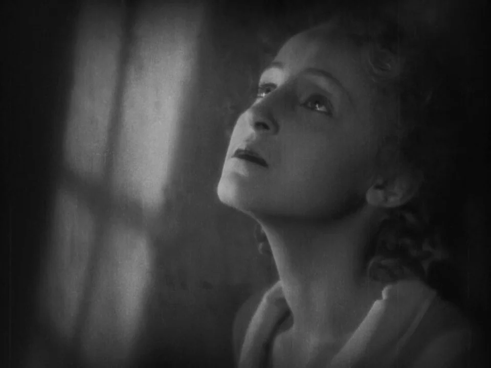 Metropolis (1927) movie still: closeup — A dimly lit interior space with a distinct shadow of a window frame. A woman gazes upward with a neutral…