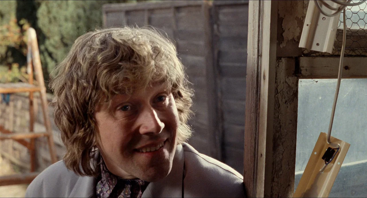 Life Is Sweet (1990) movie still: closeup — An outdoor area with a wooden fence and a window frame. A man smiles while looking at something just out…