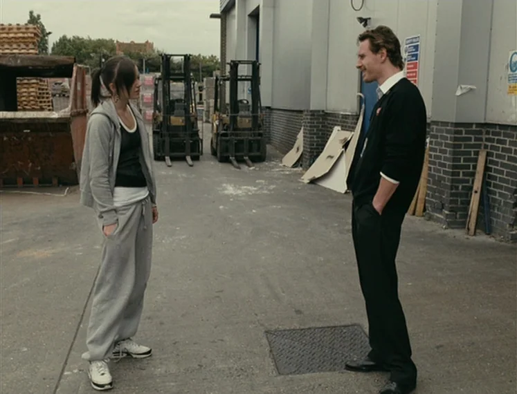 Fish Tank (2009) movie still: fullbody — A paved industrial space features two yellow forklifts and a large metal dumpster container. A woman in a…