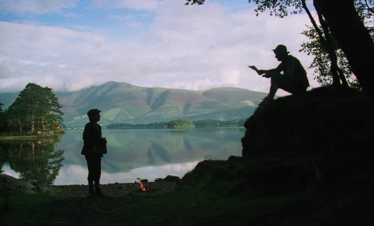 Mahler (1974) movie still: wide — Two men stand near a small campfire on the edge of a mountain lake. Mountains rise in the background with their…