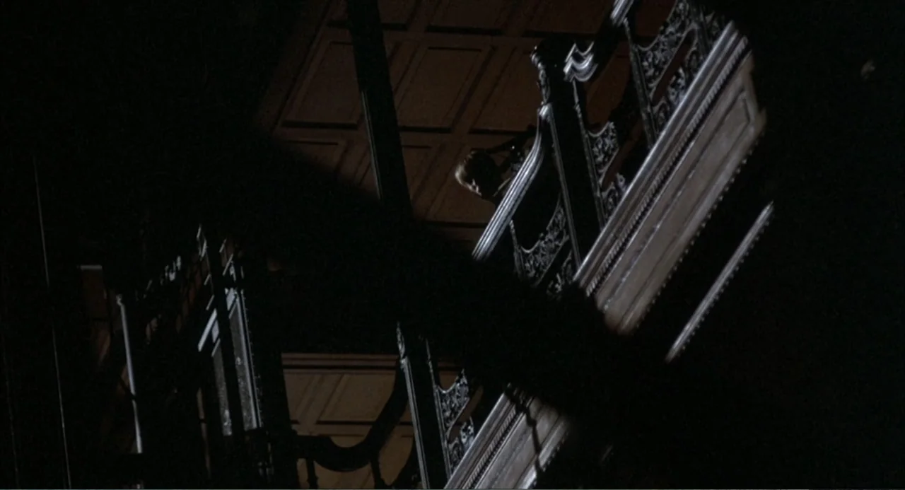 Murphy's Law (2009) movie still: wide — A dark, ornate wooden staircase structure fills the foreground and midground of the frame. A lone person…