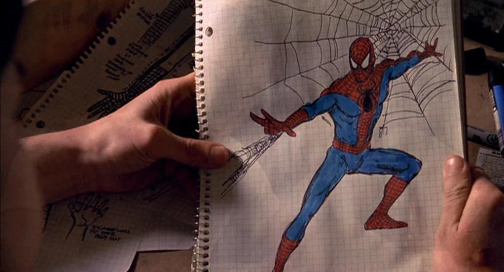 Spider-Man (2002) movie still: closeup — Hands hold open a spiral-bound notebook containing a hand-drawn sketch. The drawing depicts a costumed…