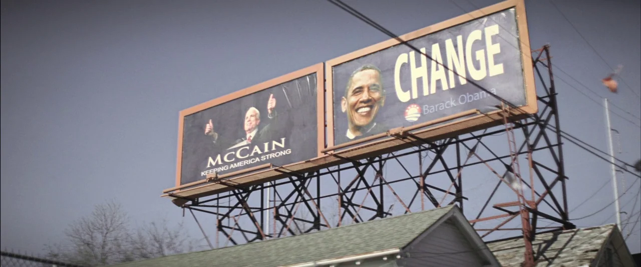 Killing Them Softly (2012) movie still: wide — Large double billboards sit atop a metal support structure against a clear sky. The placards depict…