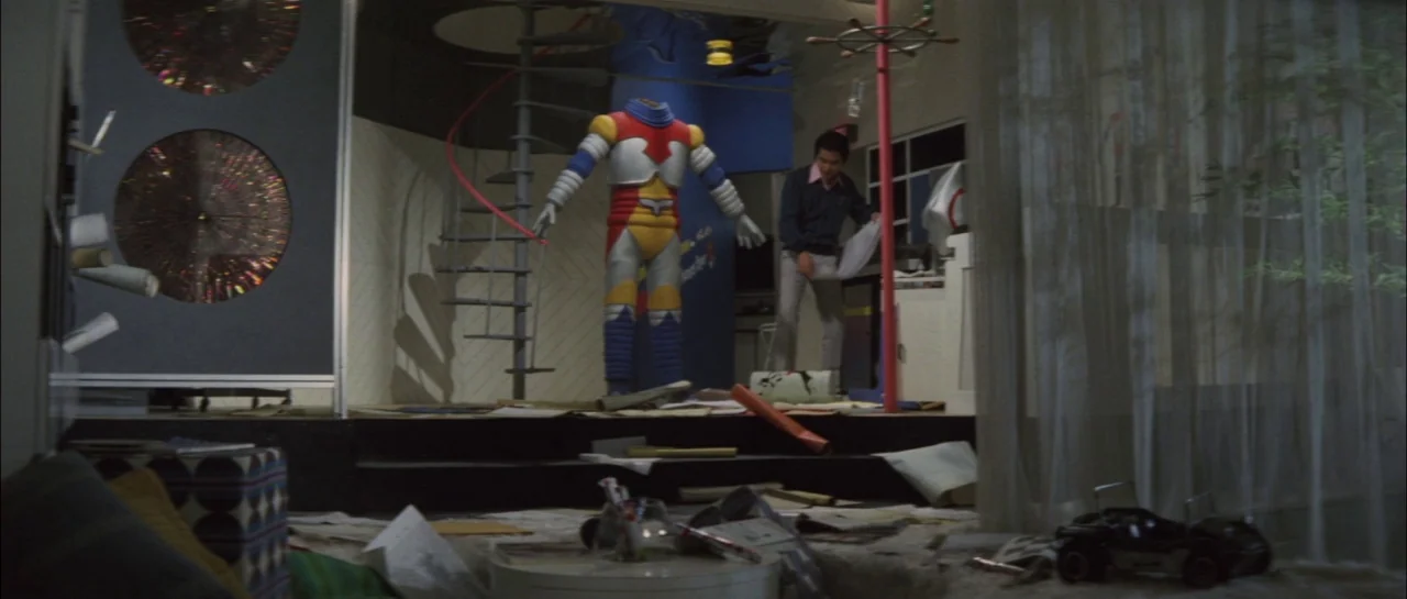 Godzilla Vs Megalon (1973) movie still: wide — A cluttered apartment room features a headless robot suit displayed near a spiral staircase. A man…