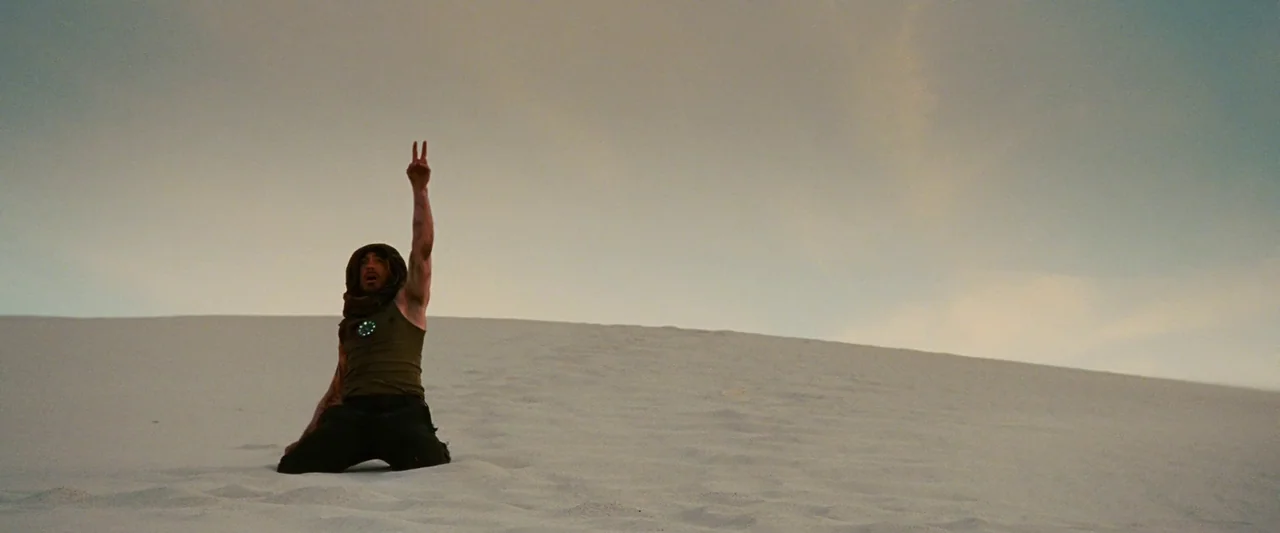 Iron Man (2008) movie still: wide — A vast expanse of white sand dunes under an overcast, hazy sky. A lone man kneels in the center sand, gesturing…