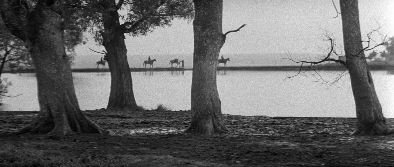 Andrei Rublev (1966) movie still: wide — A dark muddy shoreline is framed by several thick tree trunks in the foreground. Four figures on horseback…