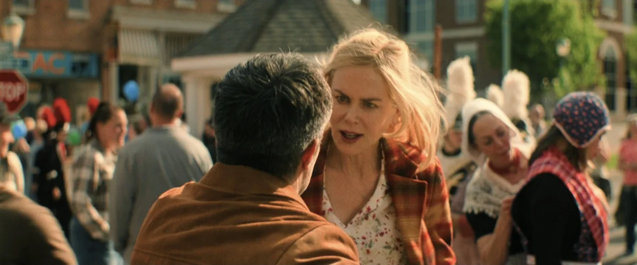 Holland (2025) movie still: medium — A public square during a community event with blurred spectators and brick buildings visible. A woman addresses…