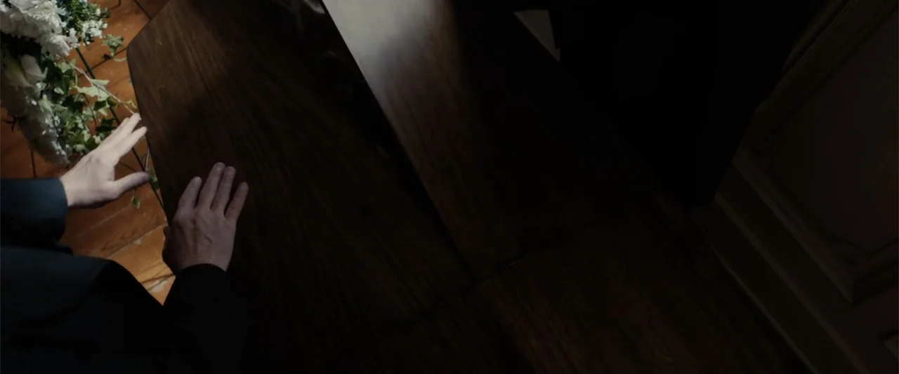 I Heard the Bells (2022) movie still: closeup — A dark wooden casket rests in a room with a hardwood floor. A person in a black suit places both…