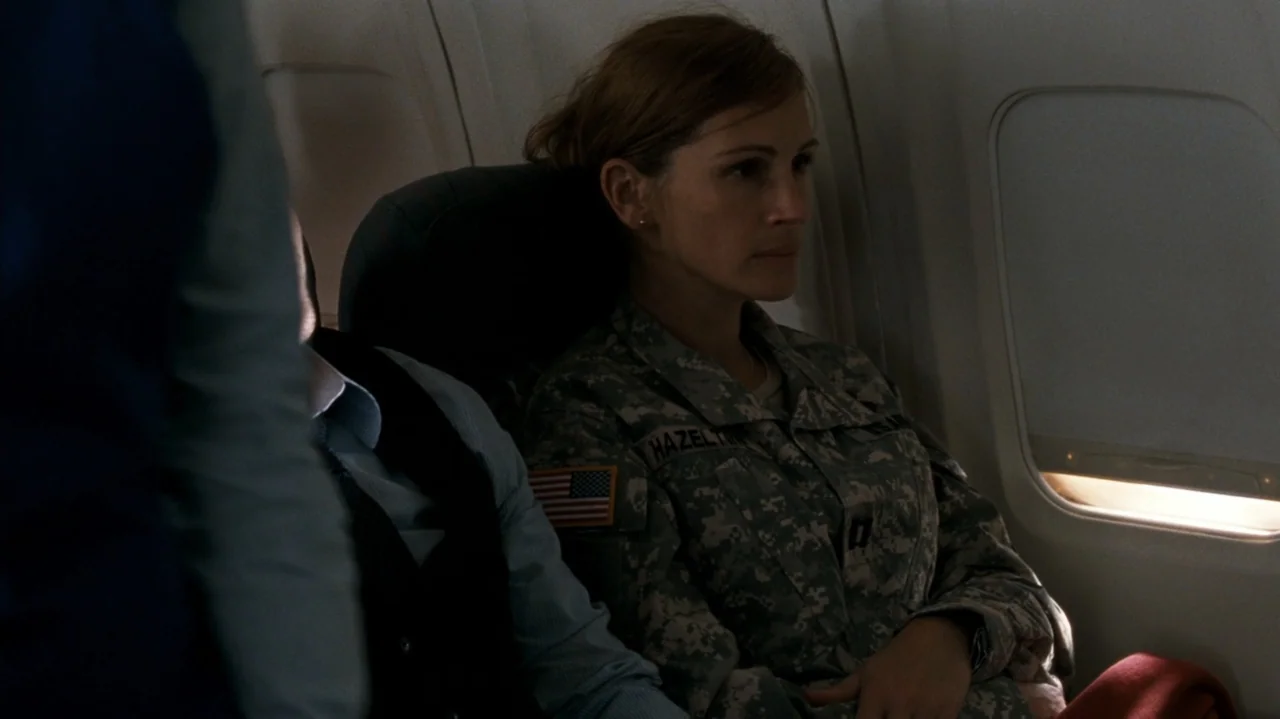F Valentine's Day (2026) movie still: medium — A woman in a military uniform sits in a cramped airplane seat near a window. She rests her hands on…