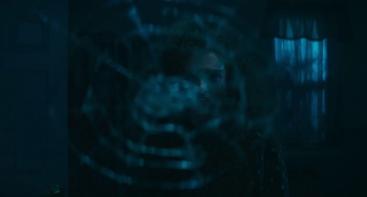 Lisa Frankenstein (2024) movie still: closeup — A dark, dimly lit room is viewed through a cracked and shattered glass pane. A woman with curly hair…