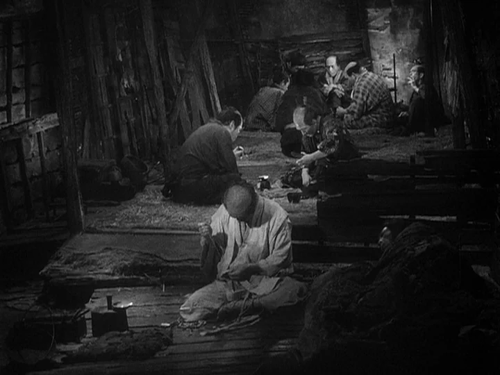 The Lower Depths (1957) movie still: wide — A dilapidated, crowded interior with wooden walls and floor mats where people reside. Several men are…
