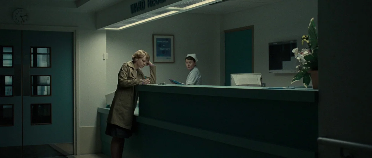 Never Let Me Go (2010) movie still: wide — A quiet hospital reception desk with teal surfaces and dull, clinical interior walls. The woman stands at…
