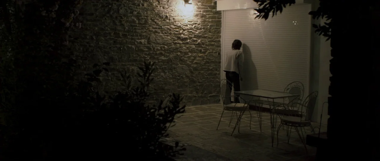 Alps (2011) movie still: wide — A stone-walled patio at night contains a glass-topped table and metal chairs. A woman stands in front of a white…