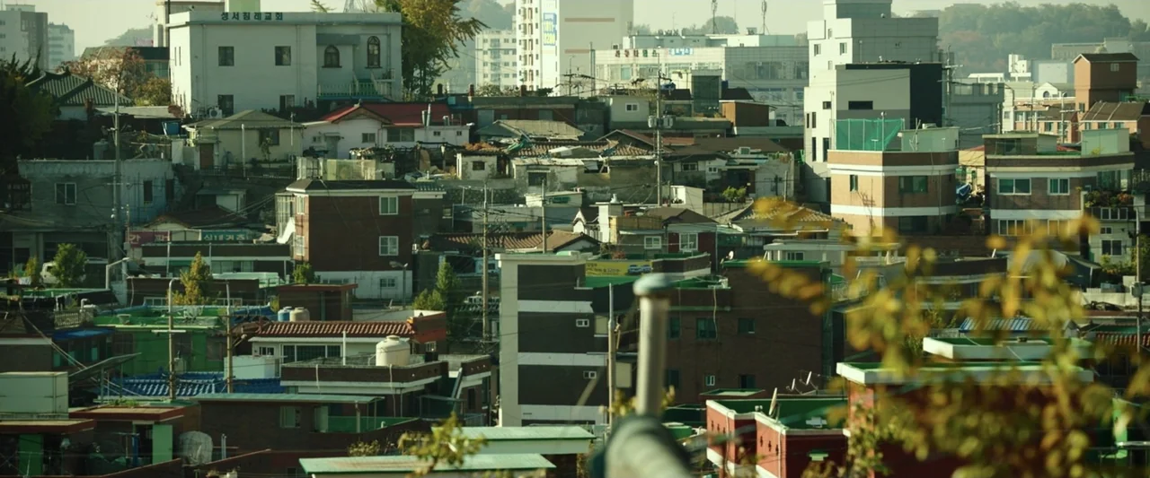 The Gangster, the Cop, the Devil (2019) movie still: establishing — A dense urban residential neighborhood features low-rise buildings and power…