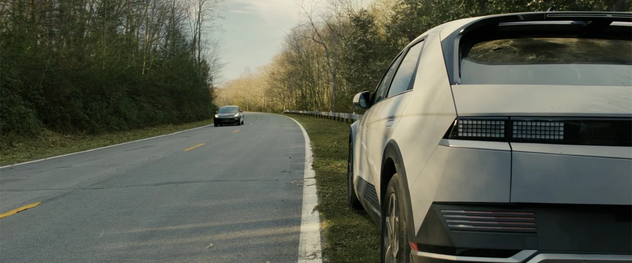 Companion (2025) movie still: medium — A rural asphalt road cuts through a dense forest with bare trees during daytime. A silver hatchback car sits…