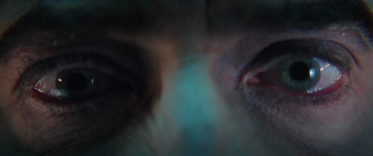 Renfield (2023) movie still: closeup — A close-up view focuses on the eyes of a person in a dark space. The skin texture is visible while a teal…