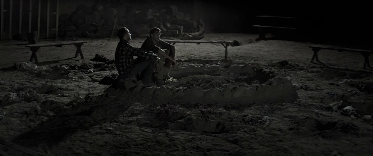 The Endless (2017) movie still: wide — An outdoor campsite at night features a ring of sand and stones. Two men sit on the edge of the pit in…