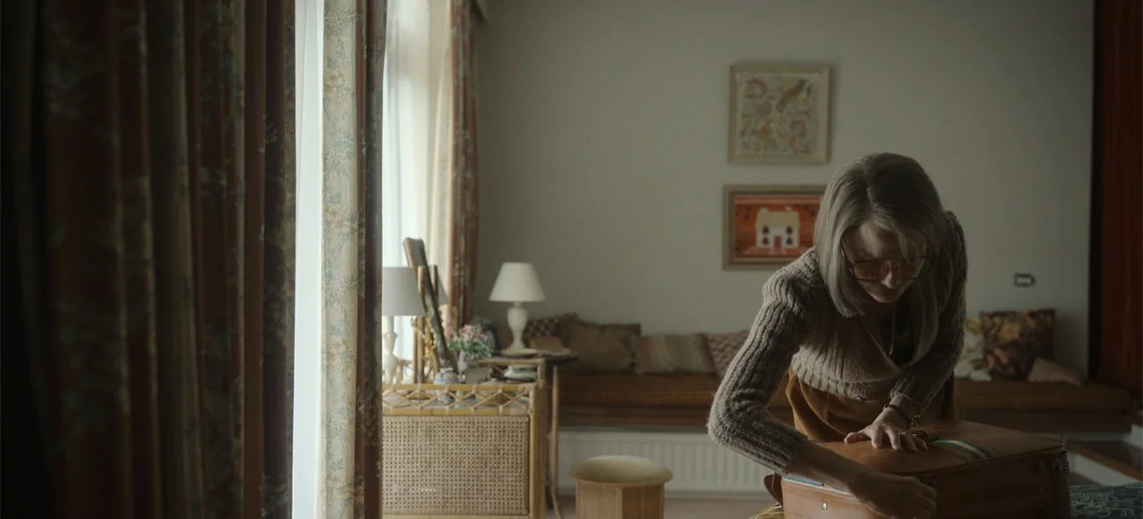 One Life (2023) movie still: medium — A mid-century style living room with patterned curtains, rattan furniture, and framed wall art. A woman leans…