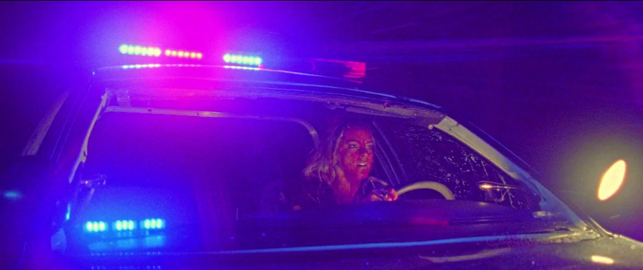 Christmas Bloody Christmas (2022) movie still: medium — A woman sits inside a police cruiser at night under flashing red and blue lights. The car…