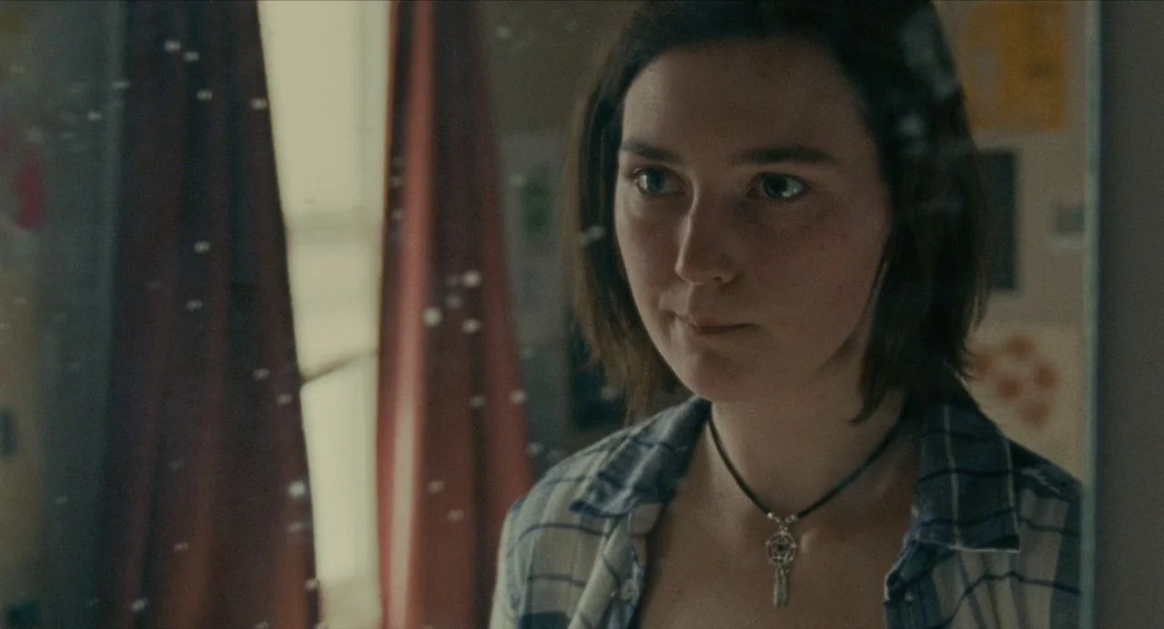 Never Rarely Sometimes Always (2020) movie still: closeup — A person stands in front of a mirror with a window and curtains visible behind. A woman…