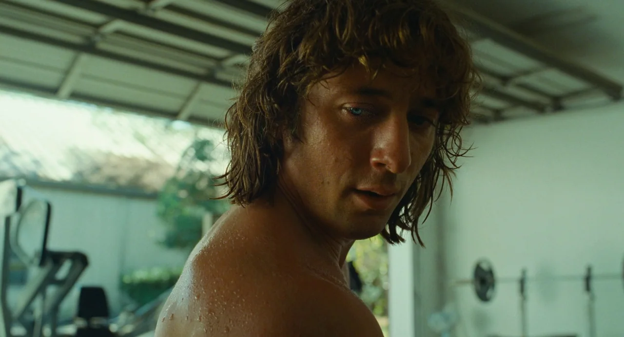 The Iron Claw (2023) movie still: closeup — A garage with workout equipment in the background is lit by soft daylight. A sweaty man with wavy brown…