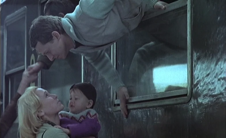 Blind Chance (1987) movie still: medium — A passenger train car exterior made of dark weathered metal stands at a platform. A man reaches out of a…