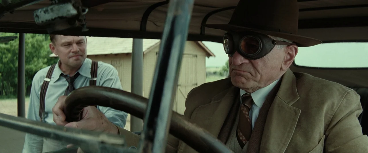 Killers of the Flower Moon (2023) movie still: medium — Two men sit inside an open vintage vehicle on a rural path. The driver wears leather goggles…