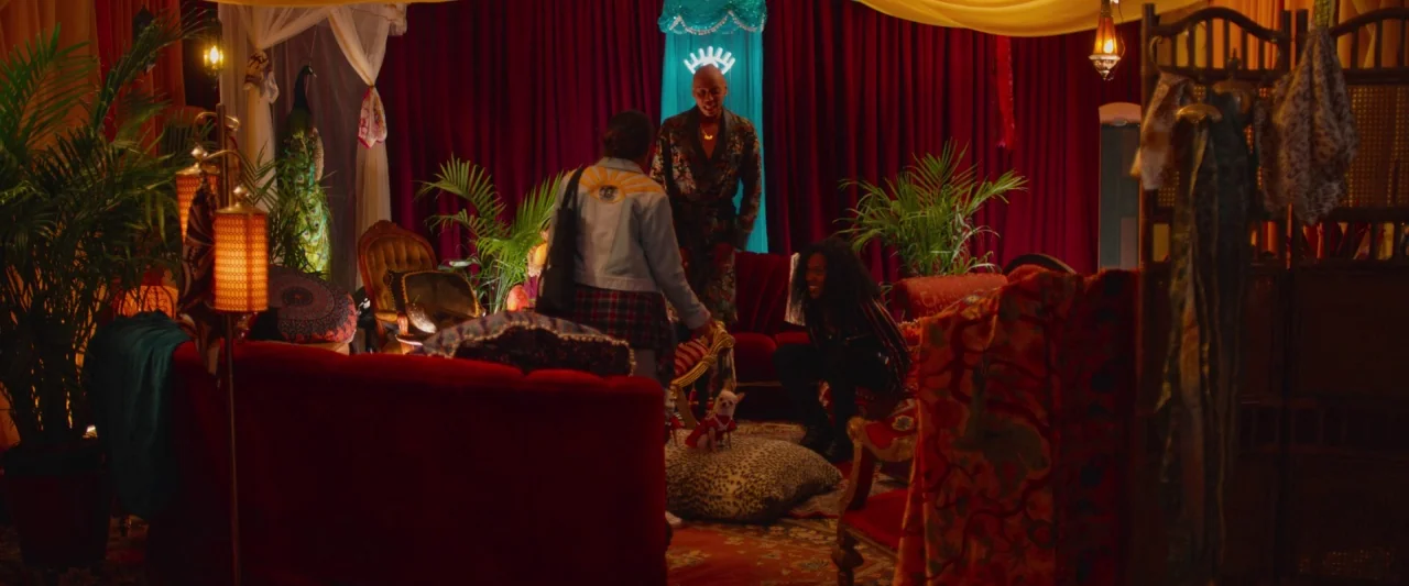 Someone Great (2019) movie still: wide — A room features heavy red velvet curtains, lush tropical plants, and eclectic vintage furniture. Three…