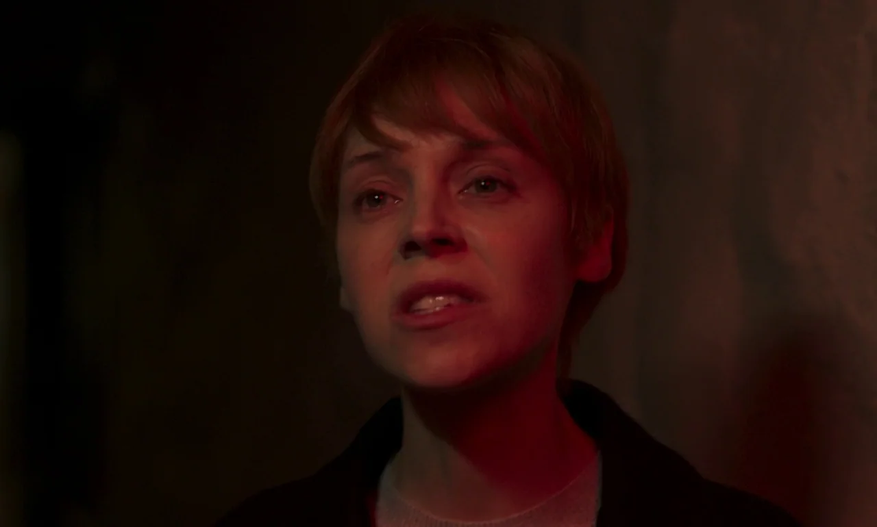It Is In Us All (2022) movie still: closeup — A dark indoor space with minimal background detail visible in low light. A woman with short hair…