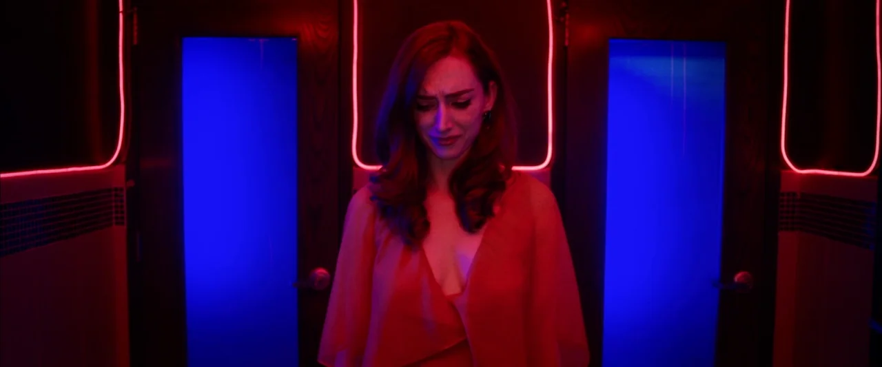The Stylist (2020) movie still: medium — A restroom stall entrance features dark wood doors outlined in glowing red neon tubing. A woman stands…