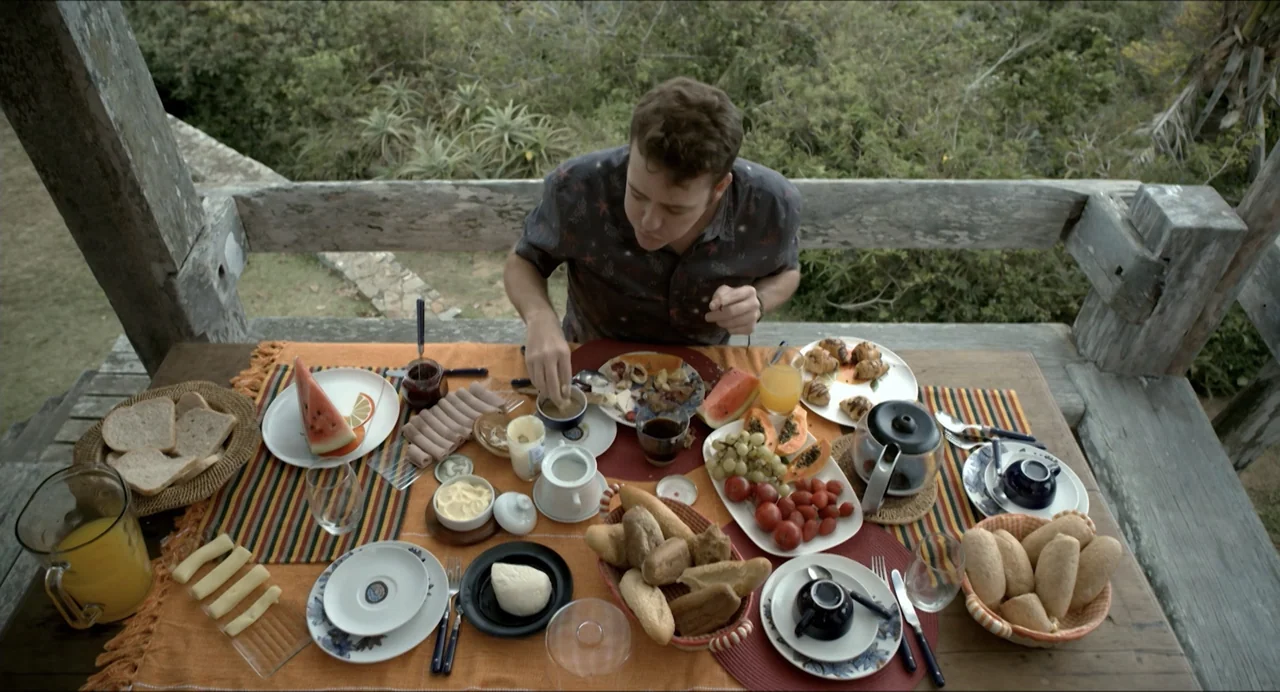 Suspension Points (2022) movie still: medium — A wooden balcony overlooks a lush tropical forest with a heavily laden breakfast table. A man sits at…