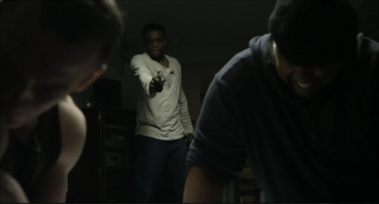 Freedom (2024) movie still: medium — A dimly lit room with an obscured background and three figures in the foreground. A man in the center holds a…