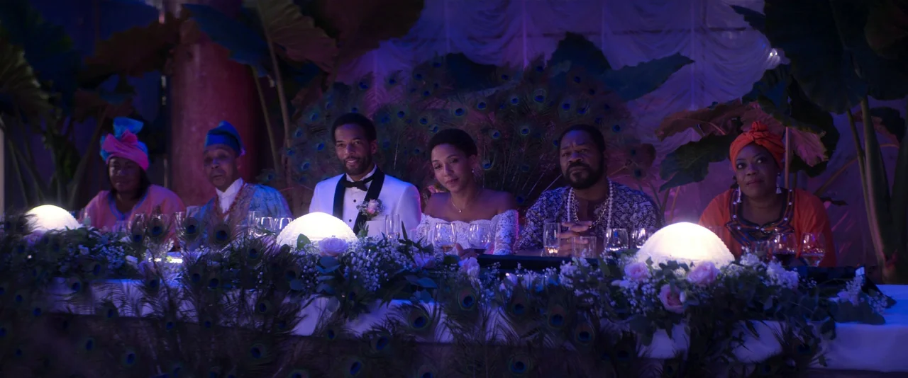 Crazy About Her (2021) movie still: medium — A wedding head table is decorated with peacock feathers, tropical leaves, and glowing globes. Six…