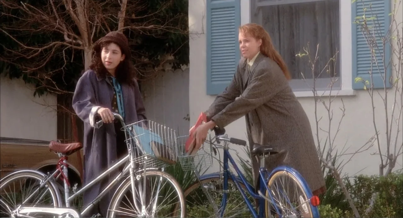 Teen Witch (1989) movie still: fullbody — A residential house front with light-colored siding and blue shutters during the day. Two young women…