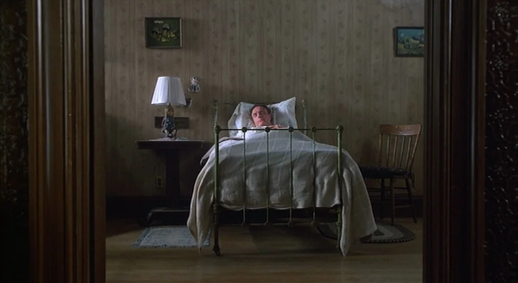 Misery (1990) movie still: wide — A sparsely furnished room features floral wallpaper and antique wood-framed doorways. A man rests in a metal bed…