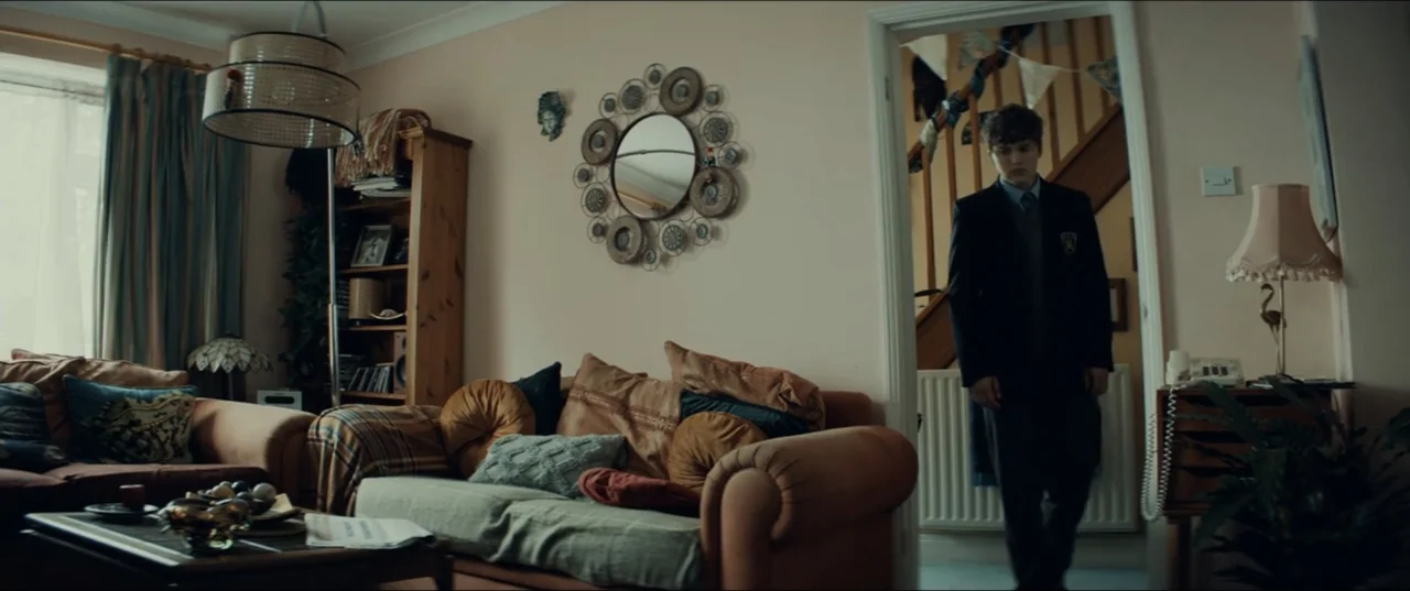 Bonus Track (2024) movie still: medium — A residential living room contains an orange couch and wall-mounted decorative circular mirrors. A teenage…