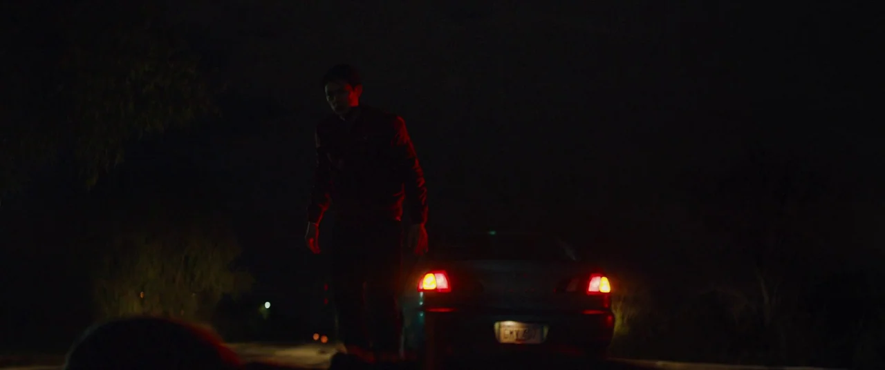 Broadcast Signal Intrusion (2021) movie still: fullbody — A dark, unlit road at night with minimal surrounding terrain visible. A man stands near…