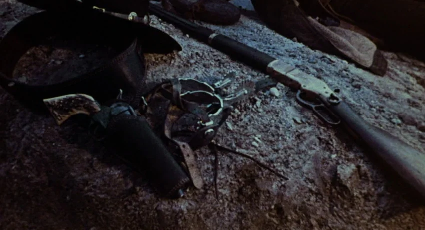 Roy Colt and Winchester Jack (1970) movie still: closeup — A collection of 19th-century frontier gear lies scattered on a coarse, dirt surface. A…