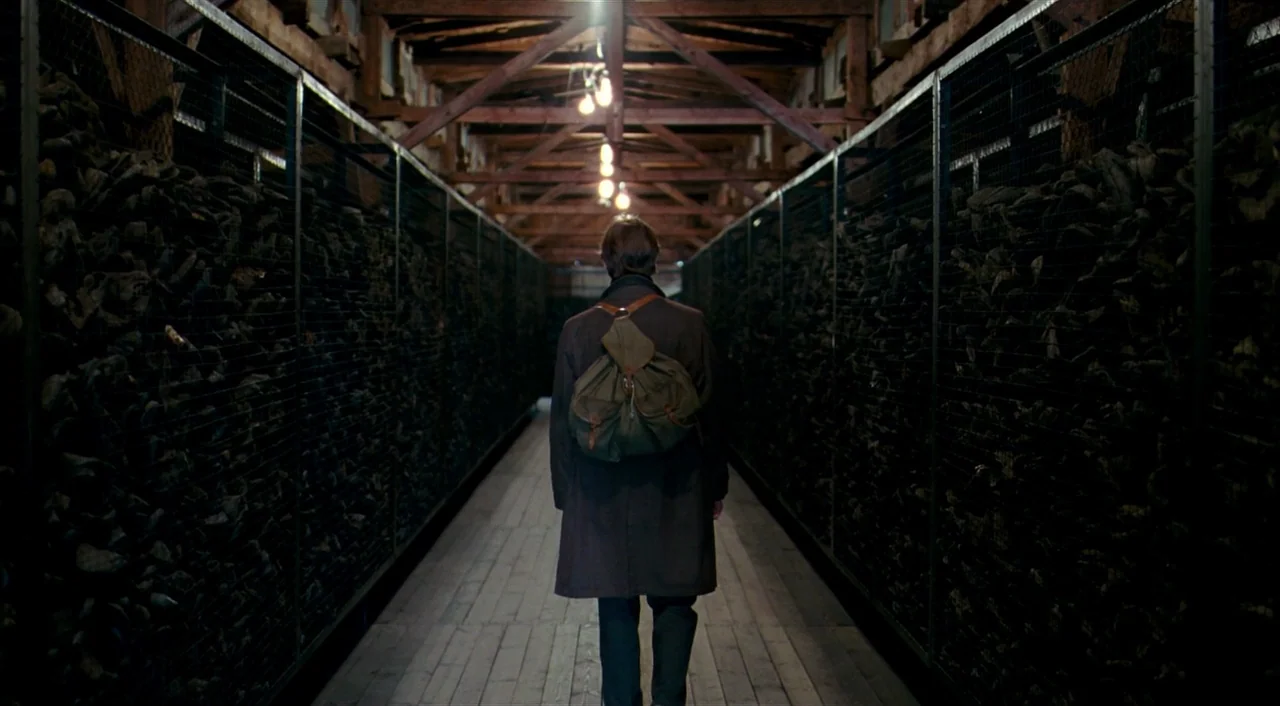 The Reader (2008) movie still: fullbody — A long, narrow wooden walkway is flanked by high chain-link partitions packed with stacked debris. A man…