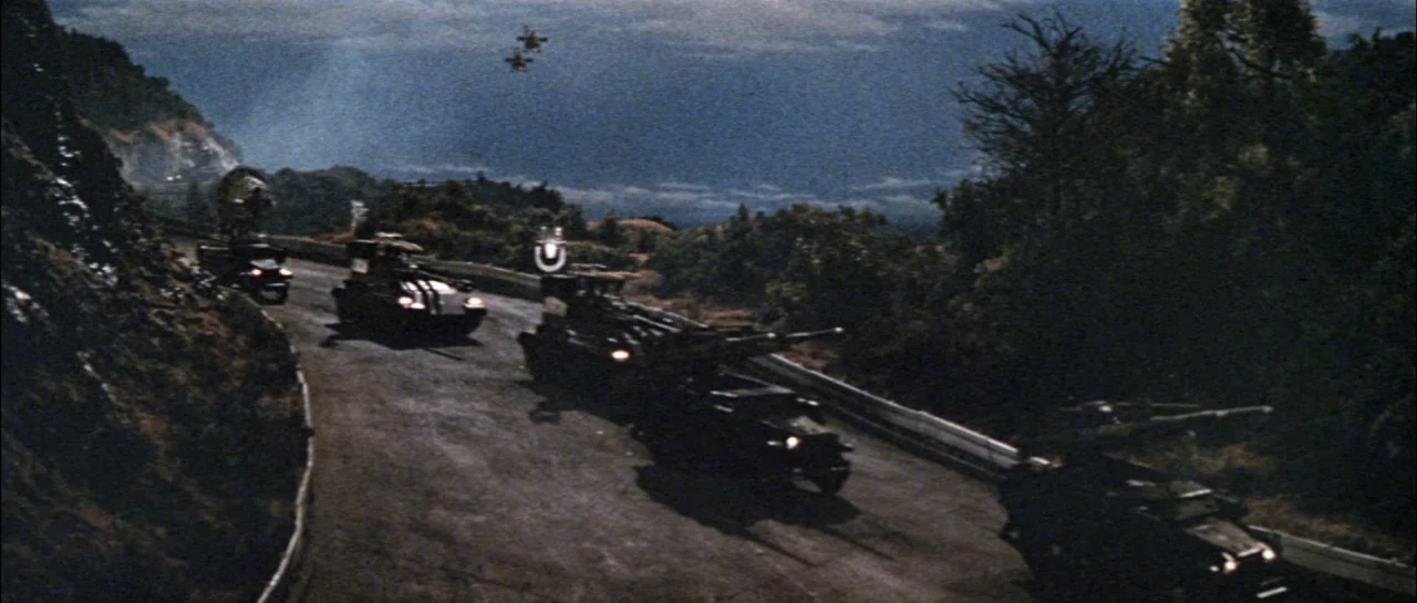Godzilla Vs Megalon (1973) movie still: wide — A dark asphalt road winds through a hilly, forested landscape under a cloudy sky. A convoy of…