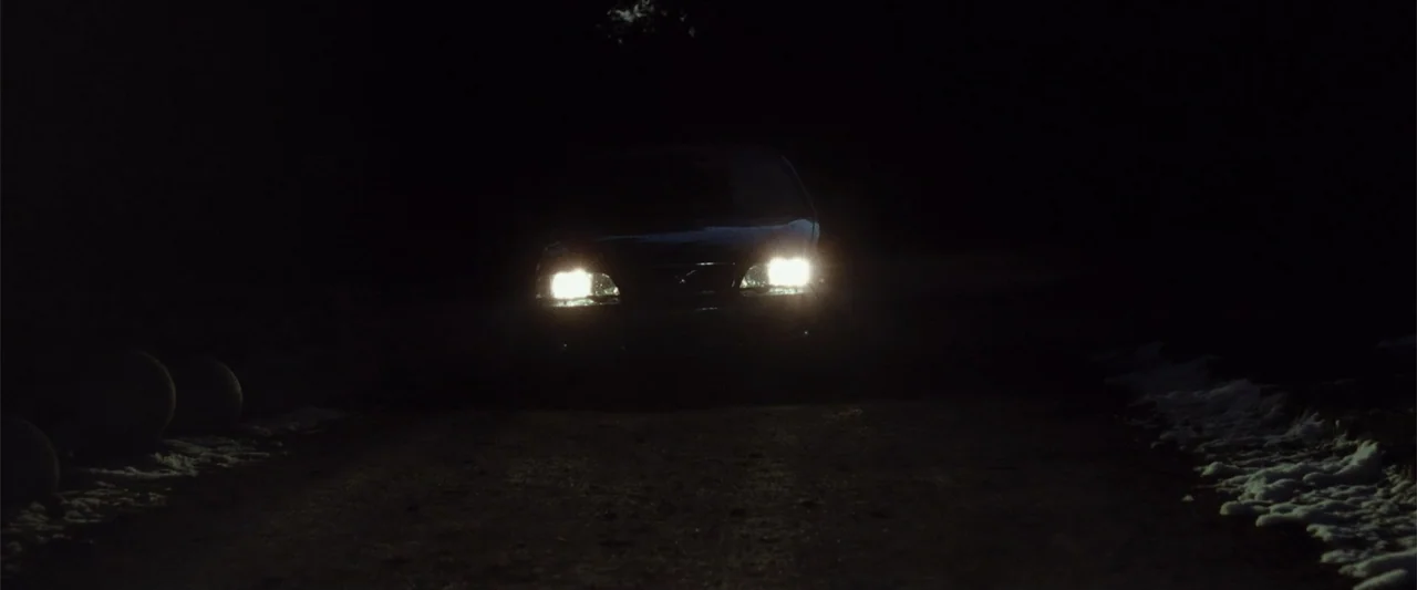 Anna (2013) movie still: wide — A dark unpaved surface stretches out into a pitch-black environment at night. A car stands centered in the distance…