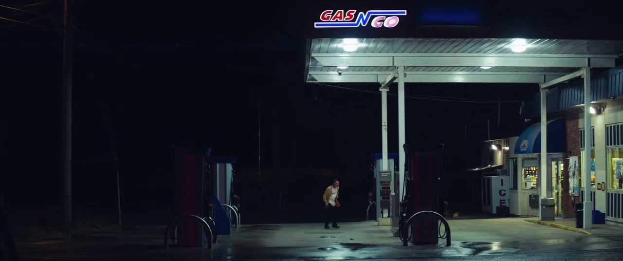 Come To Daddy (2019) movie still: wide — An illuminated gas station canopy stands against the surrounding dark night landscape. A man walks alone…