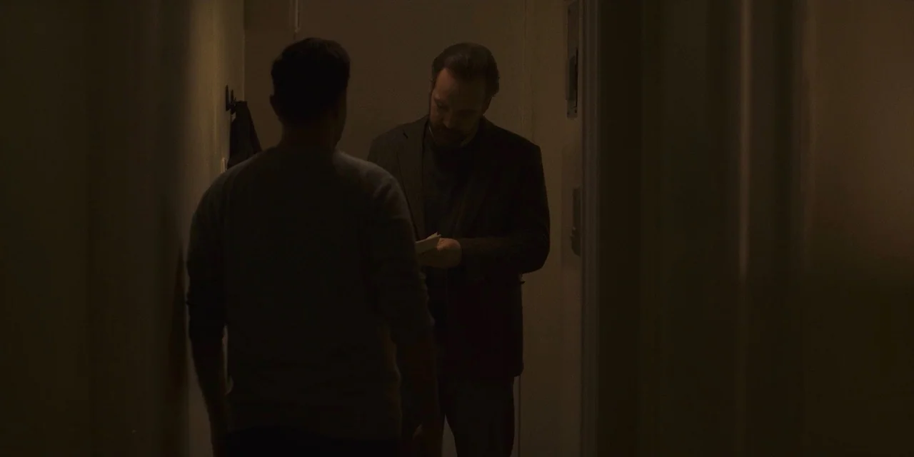 The Sound of Silence movie still: medium — A dimly lit narrow hallway with plain walls in a residential apartment building. A man in a dark blazer…